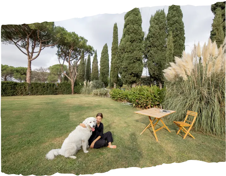 A woman sitting on the grass in a spacious backyard with a large white dog, surrounded by tall cypress trees, ornamental grass, and a wooden table with a chair, creating a serene outdoor setting.