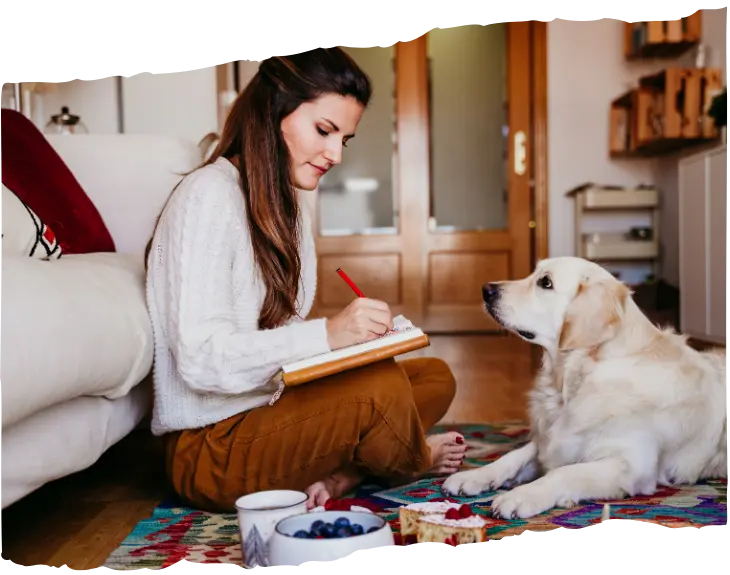 Young woman sitting on the floor, writing in a notebook while a golden retriever looks at her attentively in a cozy living room setting.